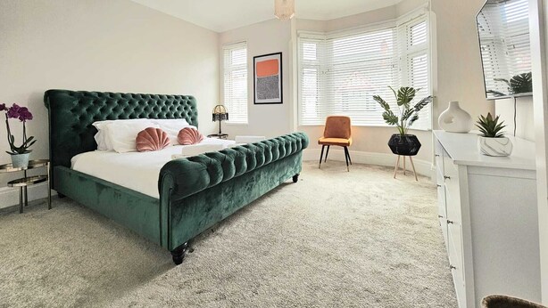A bedroom with a green upholstered bed, a small side table, a chair, a dresser, and a large window with blinds. - Home Away from Home (Coventry)