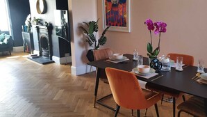 A dining area with a wooden herringbone floor, a dark wooden table set for four, and orange chairs. There is a potted plant with green leaves and purple flowers on the table. - Home Away from Home (Coventry)