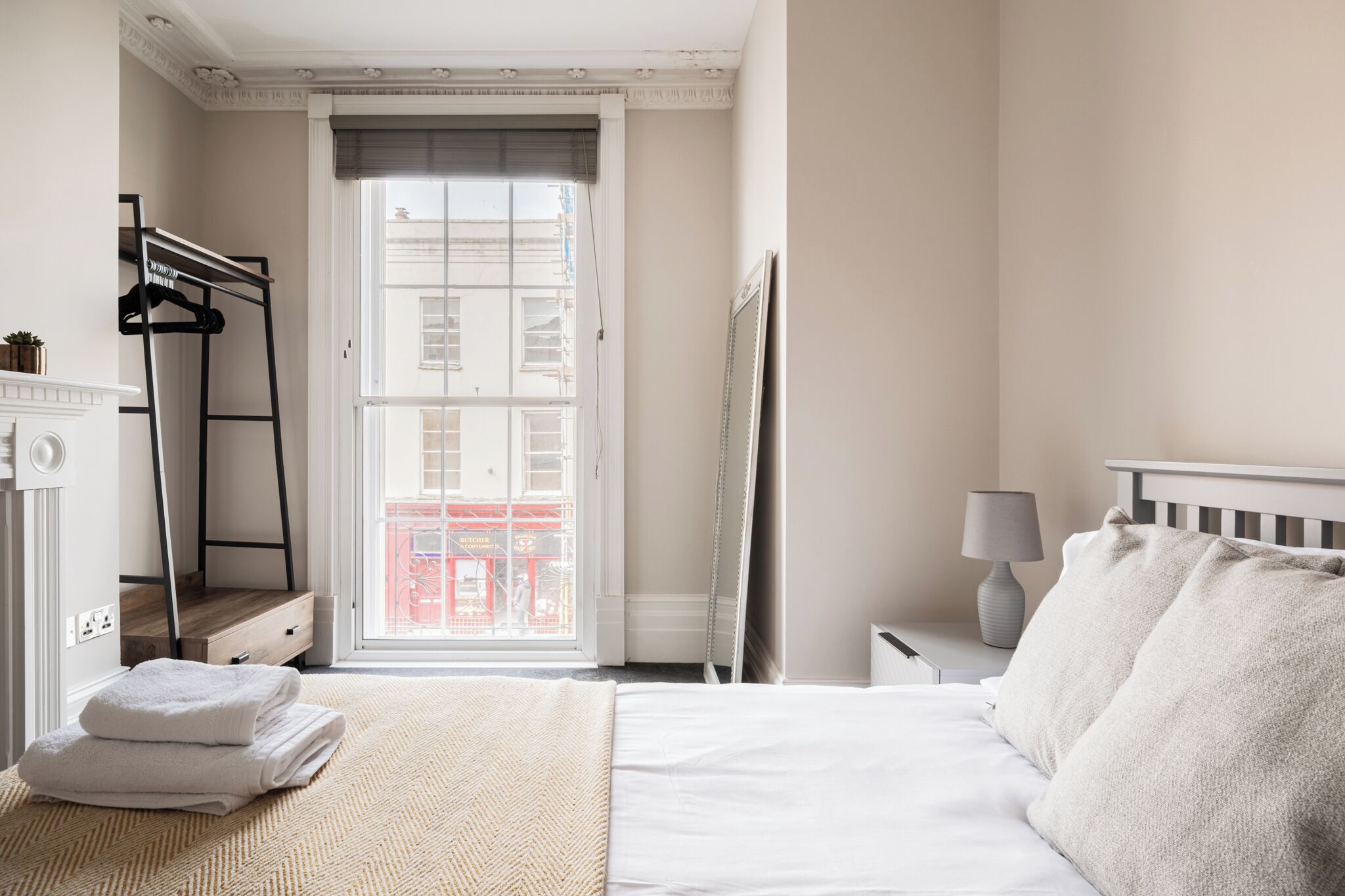 Ein Schlafzimmer mit Bett, Nachttisch mit Lampe, Fenster mit Blick auf Gebäude und Kleiderständer.