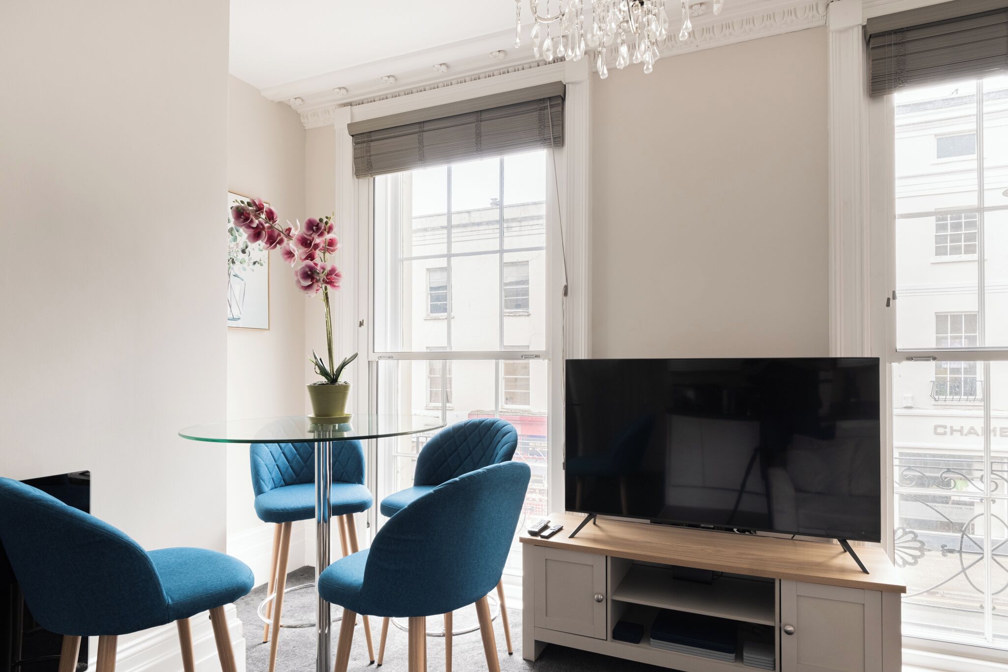 Uma sala de estar moderna com uma mesa de vidro, cadeiras azuis, uma TV de tela plana e um vaso com planta.