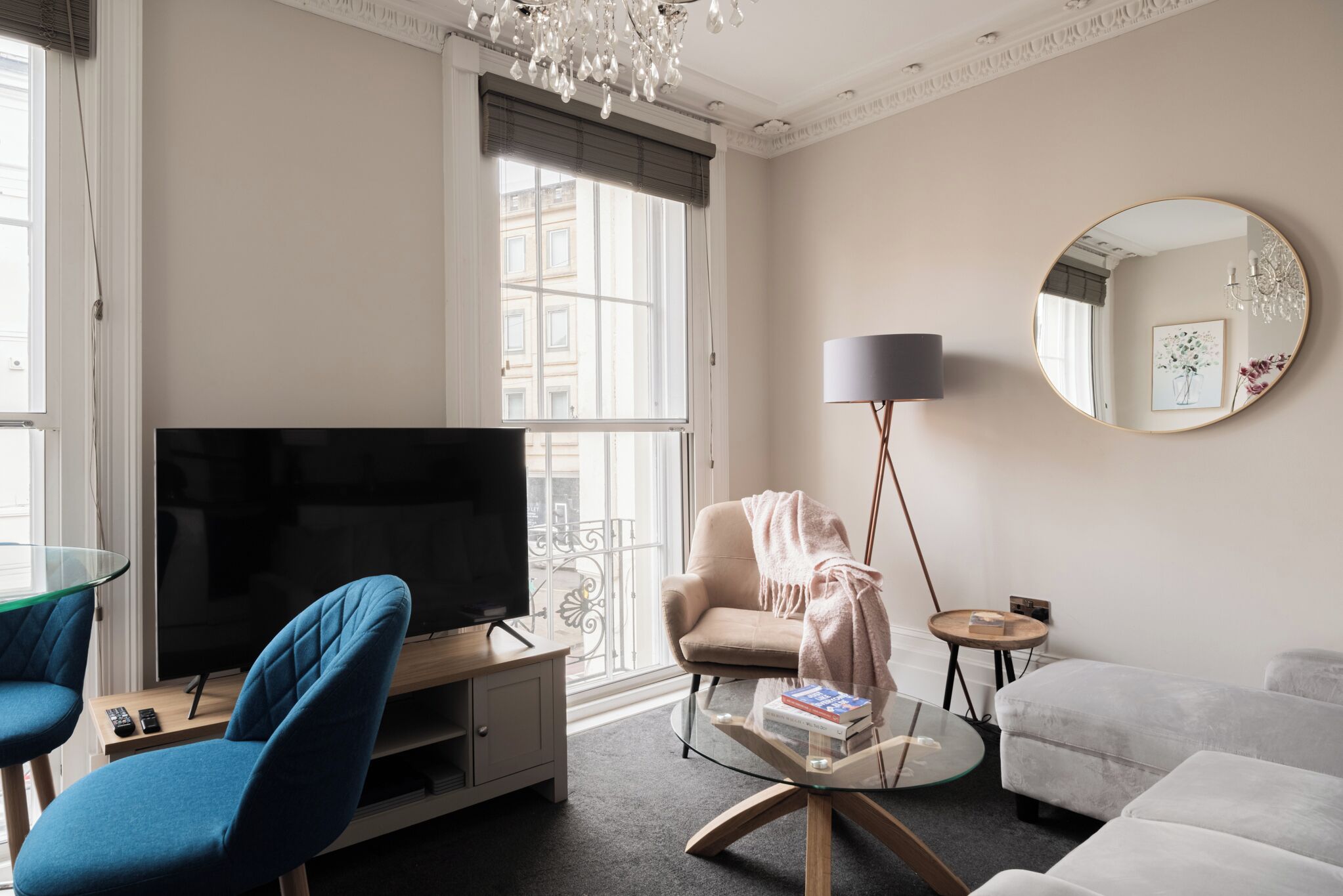 Un soggiorno moderno con una televisione a schermo piatto, un tavolino in vetro, una poltrona blu, un divano beige e uno specchio rotondo appeso al muro.