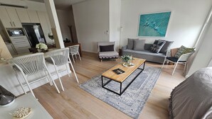 Un salon moderne avec une table basse en bois, des chaises blanches, un canapé gris et une cuisine en arrière-plan.