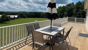 Um terraço com mesa e cadeiras, um guarda-sol preto e branco, e vista para um campo verde com árvores.