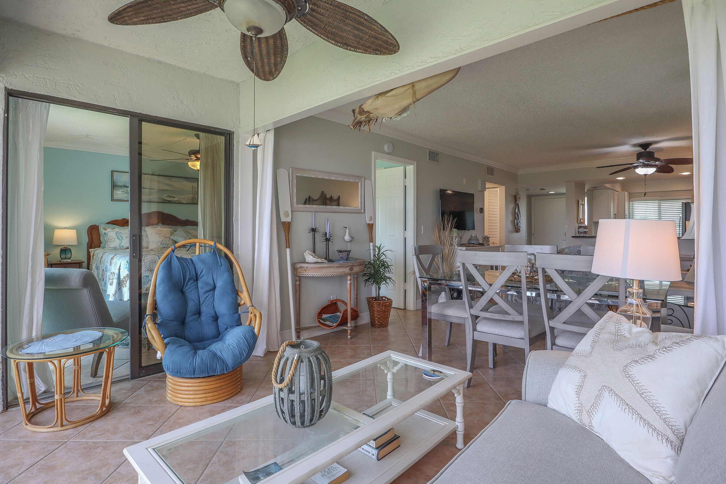 A spacious living room with a large wicker chair, a glass coffee table, and a sofa. There is a dining area with a table and chairs, and a kitchen in the background.