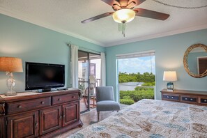 A bedroom with a bed, a television, a chair, a dresser, a ceiling fan, and a view of a lake through the window.