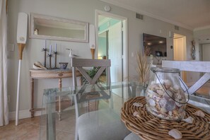 En moderne stue med et sofabord i glas, et sivbord med et glasfad fyldt med skaller, et fjernsyn og et dekorativt spejl.