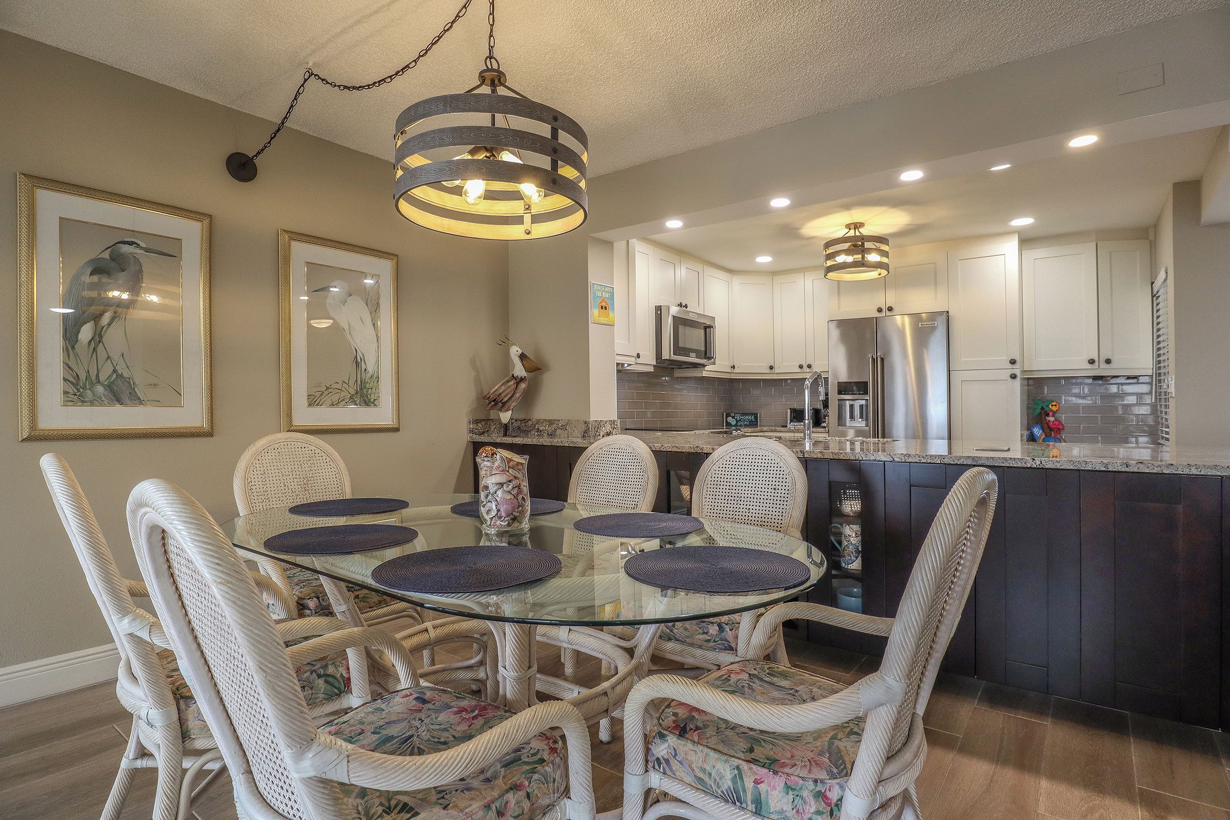 Un comedor con mesa de vidrio rodeada de sillas de mimbre, una cocina con muebles blancos y electrodomésticos de acero inoxidable, y una lámpara colgante sobre la mesa.