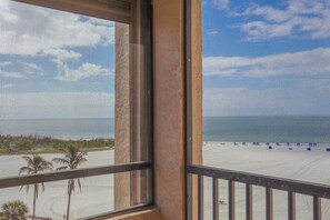 Vista desde una ventana a una playa con cielo despejado, palmeras y orilla arenosa.