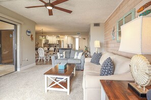 A living room with a ceiling fan, a dining area with chairs, a kitchen in the background, and a large window.