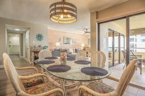 A dining area with a glass table surrounded by wicker chairs, a ceiling fan, and a view through a glass door to an outdoor balcony.
