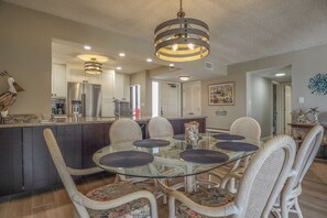 A modern kitchen with a glass dining table surrounded by wicker chairs, a stainless steel refrigerator, and a hanging light fixture.