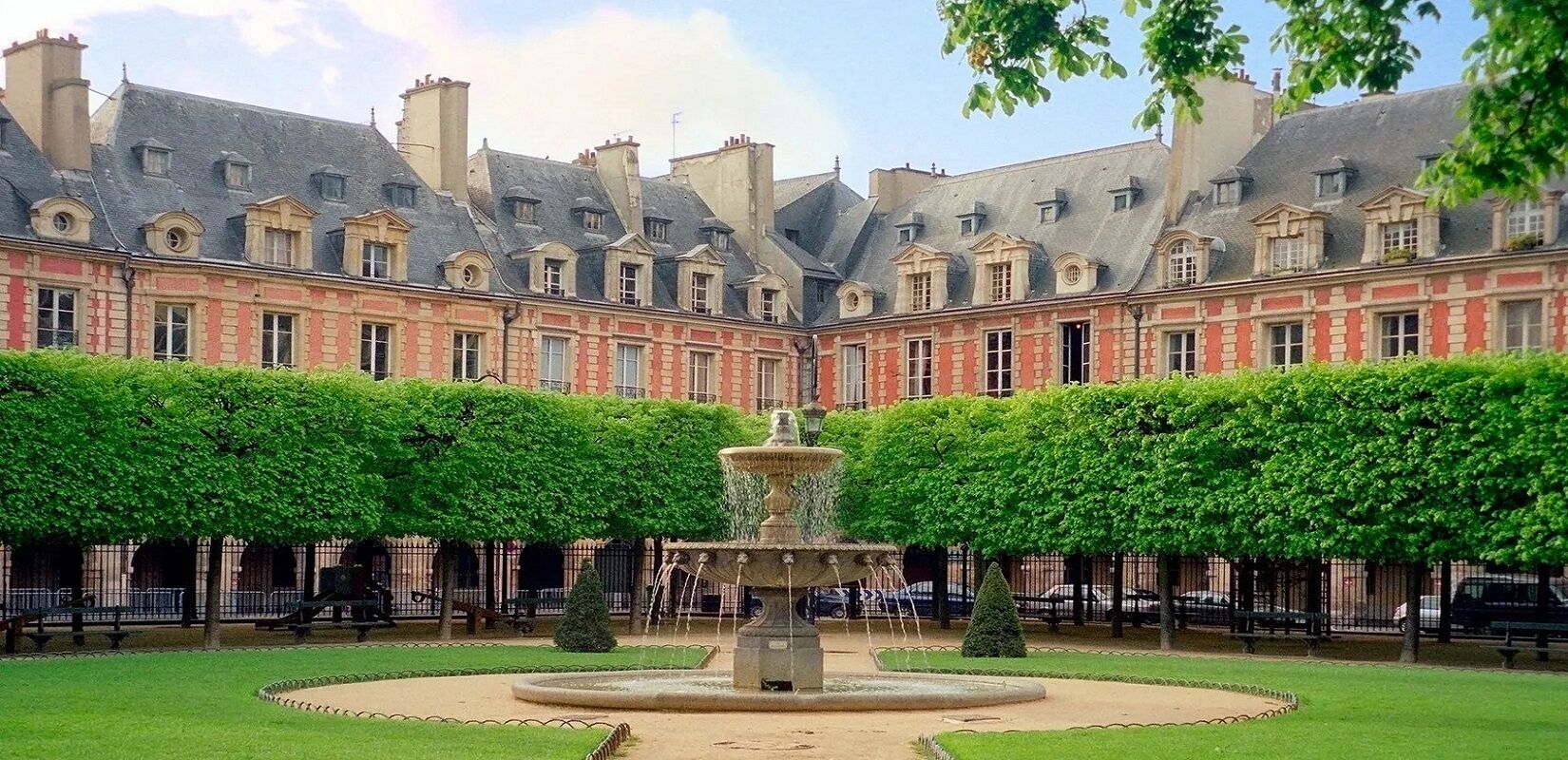 A courtyard with a fountain, surrounded by a building with red brick and grey roofs.
