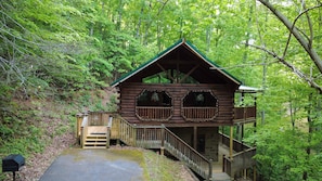 Eine Holzhütte mit Balkon, Veranda und einer Treppe, die hinauf führt, umgeben von dichtem Wald.