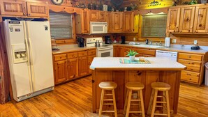 A kitchen with wooden cabinets, a refrigerator, a microwave, a stove, and a sink. There are three wooden stools at the island.