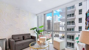A modern living room with a sofa, a glass coffee table, a potted plant, and a view of a cityscape through large windows.