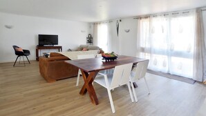 A spacious living room with a wooden dining table, white chairs, a brown sofa, a TV, and a large window with sheer curtains. - Large villa for 10 people, green setting (Férolles-Attilly)