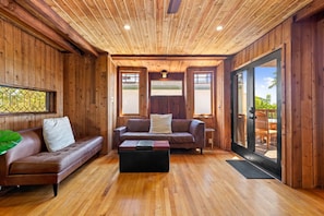 A wooden interior with a sofa, a coffee table, and a view through a glass door to an outdoor deck.