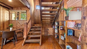 A wooden interior with a staircase, a dining table, chairs, and a bookshelf with books.