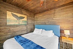 Una habitación con techo de madera, cama con ropa de cama blanca, cabecera azul, un cuadro de un pájaro y una mesita de noche con una lámpara.