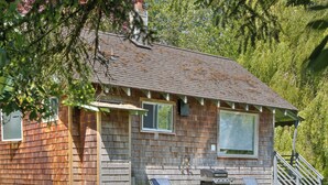 A wooden cabin with a sloped roof, two windows, a small porch with a chair, and a grill.