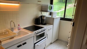 Une cuisine de taille réduite, avec des meubles blancs, un évier, une cuisinière et un four. Une fenêtre offre une vue sur la verdure environnante.