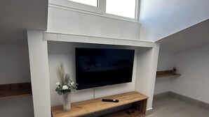 A modern living room with a flat-screen TV mounted on a white wall, wooden shelves, and a small decorative vase. - Serenity Apartments and Studios (Eforie)