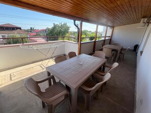 A covered balcony with a wooden ceiling, a table with wicker chairs, and a view of the street and buildings. - Serenity Apartments and Studios (Eforie)