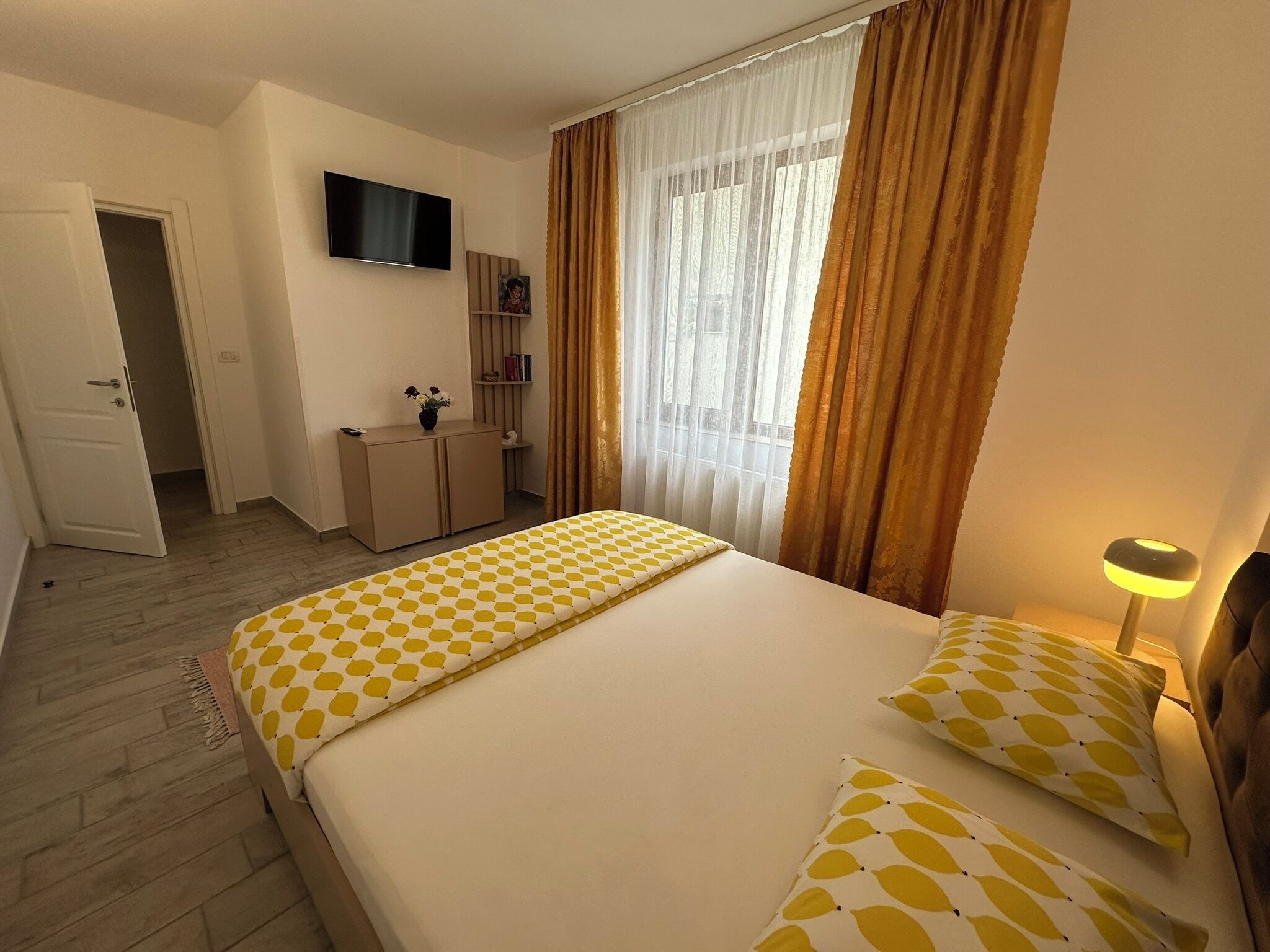 A hotel room with a bed, a TV mounted on the wall, a desk with a chair, and a window with curtains.