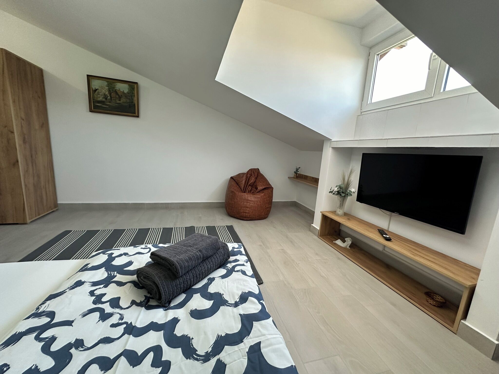 A modern bedroom with a loft-style ceiling, a large bed with patterned bedding, a bean bag chair, a flat-screen TV, and a small shelf with decorative items.
