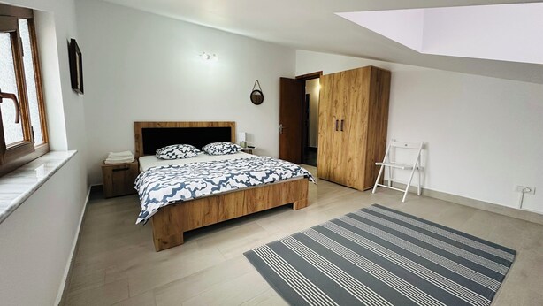 A modern bedroom with a wooden bed, a nightstand, a wardrobe, and a view of the outside through a window. - Serenity Apartments and Studios (Eforie)
