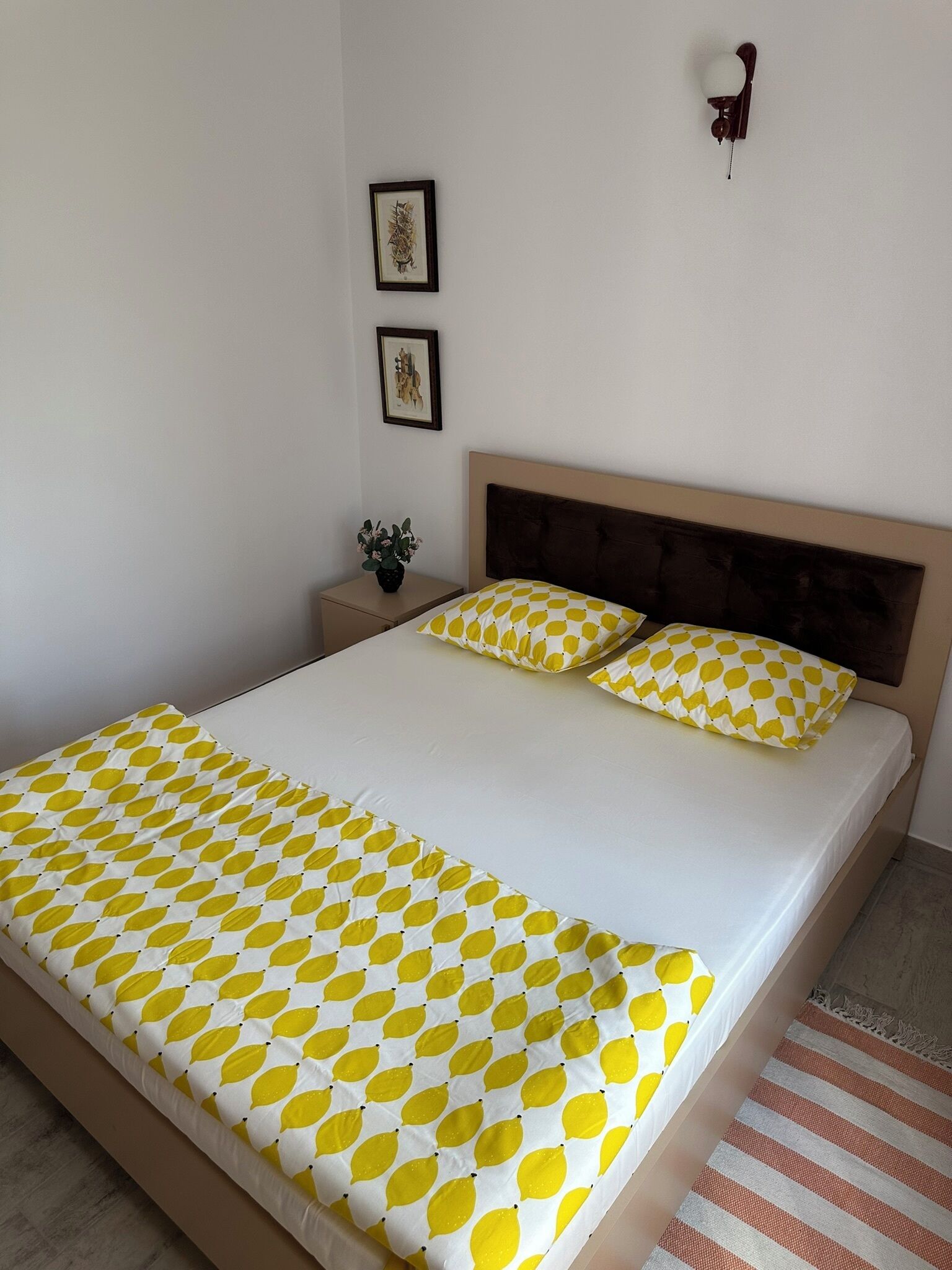 Un lit bien fait, recouvert d’une couvre-lit à motifs, une table de chevet avec une plante, et deux tableaux encadrés accrochés au mur.