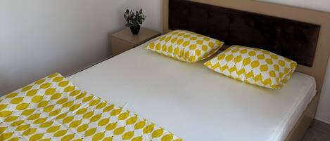 Un lit bien fait, recouvert d’une couvre-lit à motifs, une table de chevet avec une plante, et deux tableaux encadrés accrochés au mur.