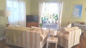 A room with striped sofas, a television, a small table with a lamp, and a view of an outdoor patio with chairs and plants. - Cosy Bayside Studio - Spring Into Relaxation By The Bay (Manly West)