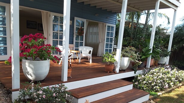 A covered porch with a wooden deck, white chairs, and potted plants. - Cosy Bayside Studio - Spring Into Relaxation By The Bay (Manly West)