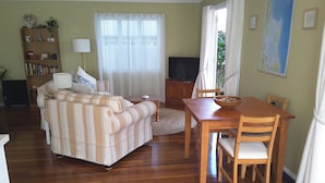 A living room with a sofa, a TV, a dining table with chairs, a bookshelf, and a window with curtains. - Cosy Bayside Studio - Spring Into Relaxation By The Bay (Manly West)