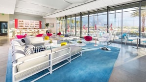 A modern lounge area with a bar, seating, and large windows offering a view of palm trees. - The Miralon Petite Oasis (Palm Springs)