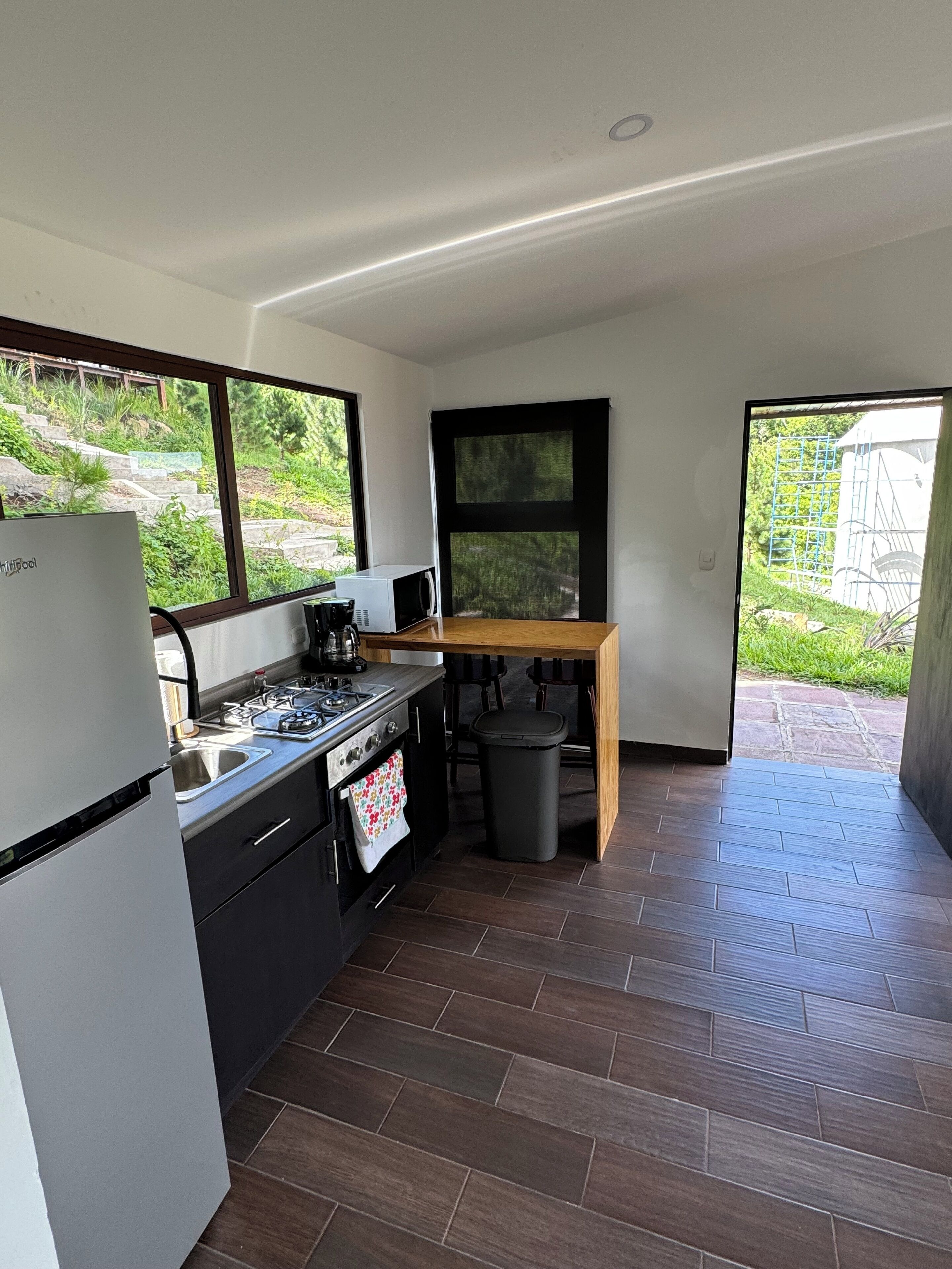 Ett modernt kök med kylskåp, spis, mikrovågsugn och diskho. Det finns ett matbord med stolar och en papperskorg. Stora fönster ger utsikt över en trädgård och en byggnad.