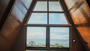 A person sitting on a bed in a room with a large window, looking out at a landscape.