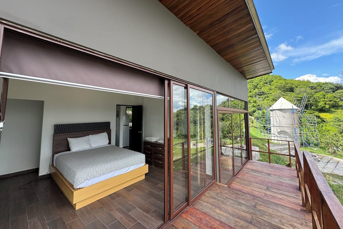A modern bedroom with a large bed, a wooden headboard, and a glass door leading to a balcony with a view of a mountain and a gazebo.