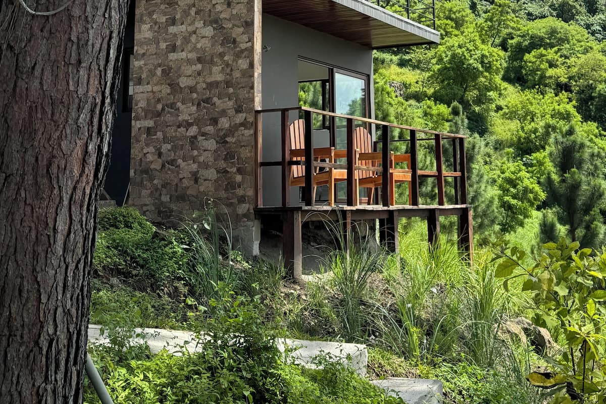 A modern two-story house with a stone facade, a balcony with wooden chairs, and a view of a lush green hillside.