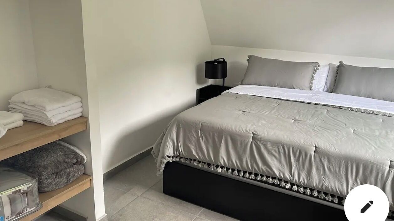 A neatly made bed with a grey duvet, a bedside table with a lamp, and a shelf with folded towels.