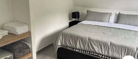 A neatly made bed with a grey duvet, a bedside table with a lamp, and a shelf with folded towels.