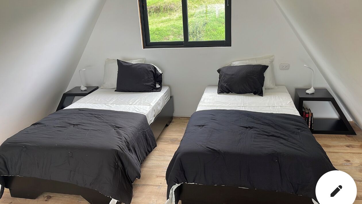 A small, modern room with two single beds, each with a nightstand and a lamp. The room has a sloped ceiling and a window providing a view of greenery outside.