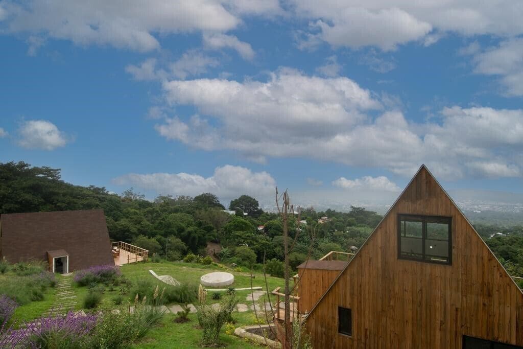 A wooden cabin with a triangular roof, surrounded by lush greenery and a clear sky.