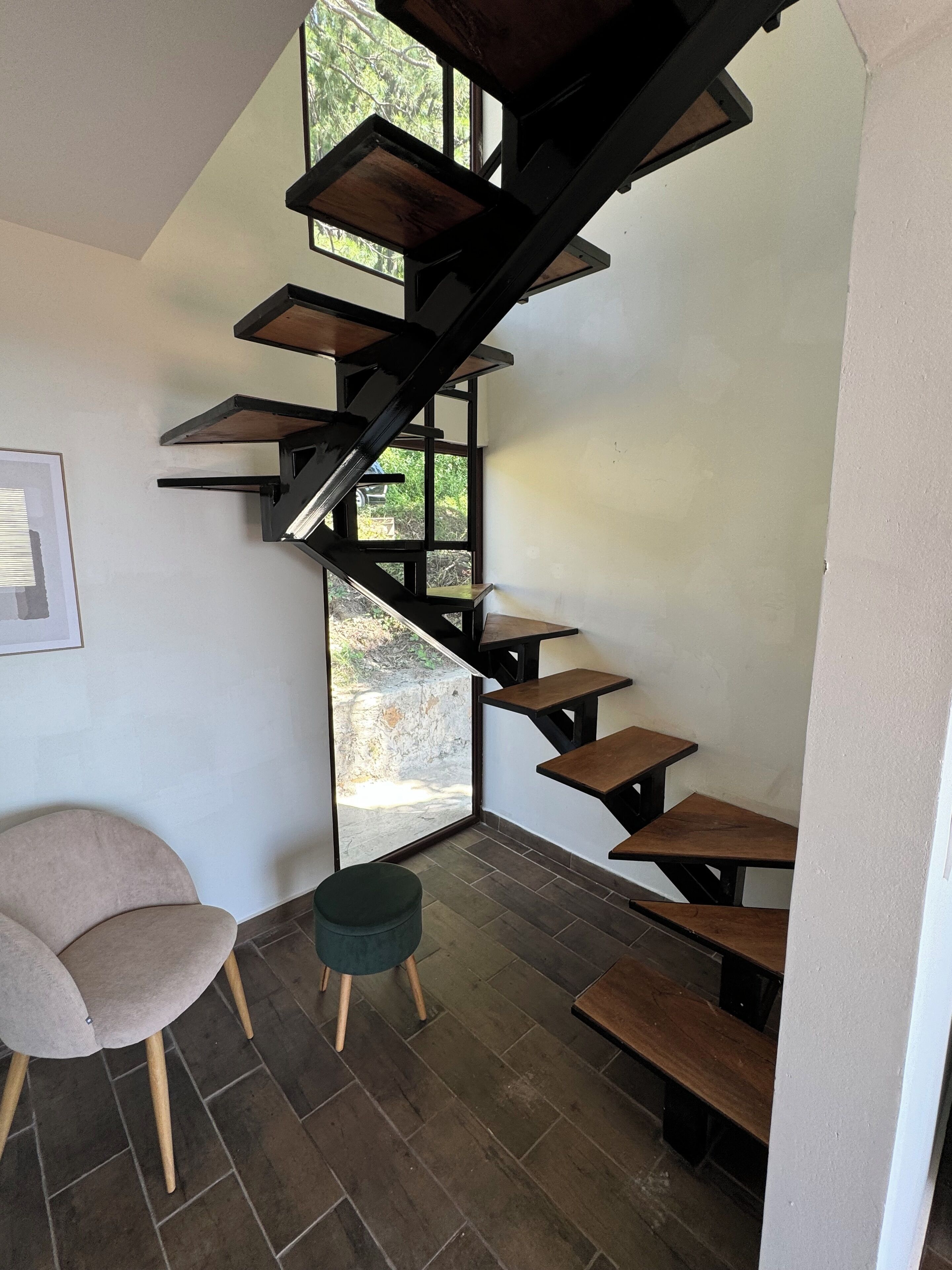 Un interior moderno con una escalera en espiral, un sillón acolchado, un taburete pequeño y un cuadro en la pared.