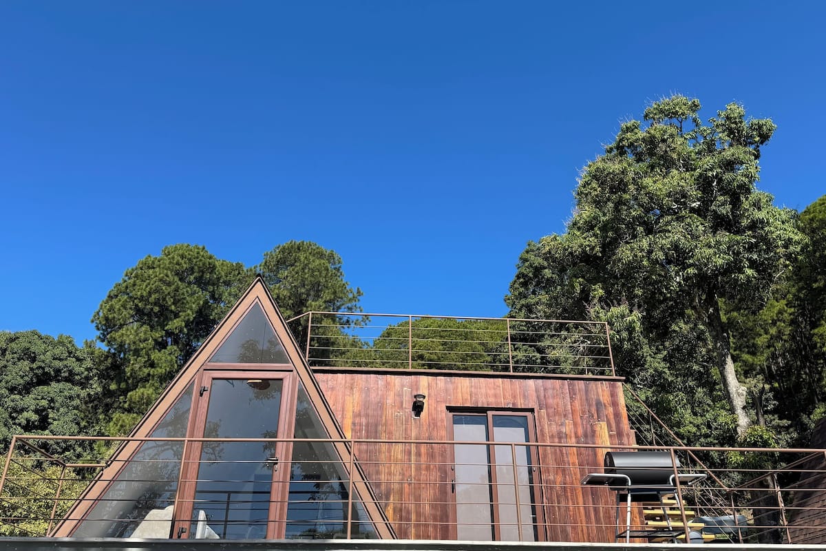 A triangular wooden cabin with a large glass window, surrounded by lush greenery and trees.