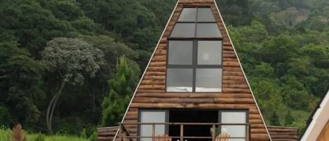 A wooden cabin with a triangular roof, large windows, and a balcony with chairs.