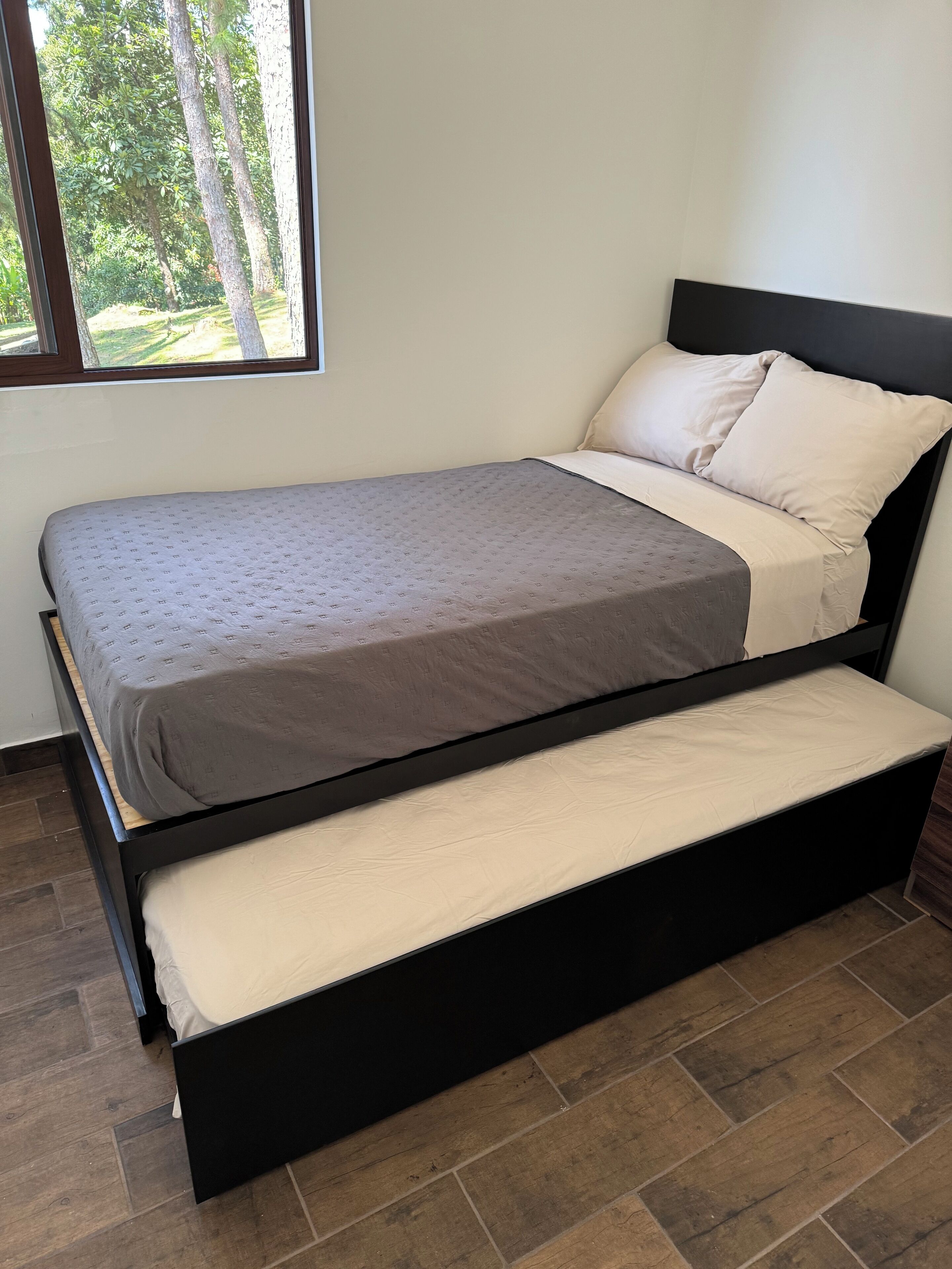 Cama doble con estructura negra, ropa de cama color beige y ventana con vista a árboles.