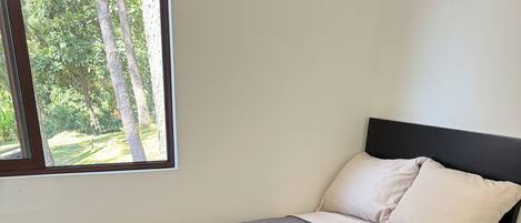 Cama doble con estructura negra, ropa de cama color beige y ventana con vista a árboles.