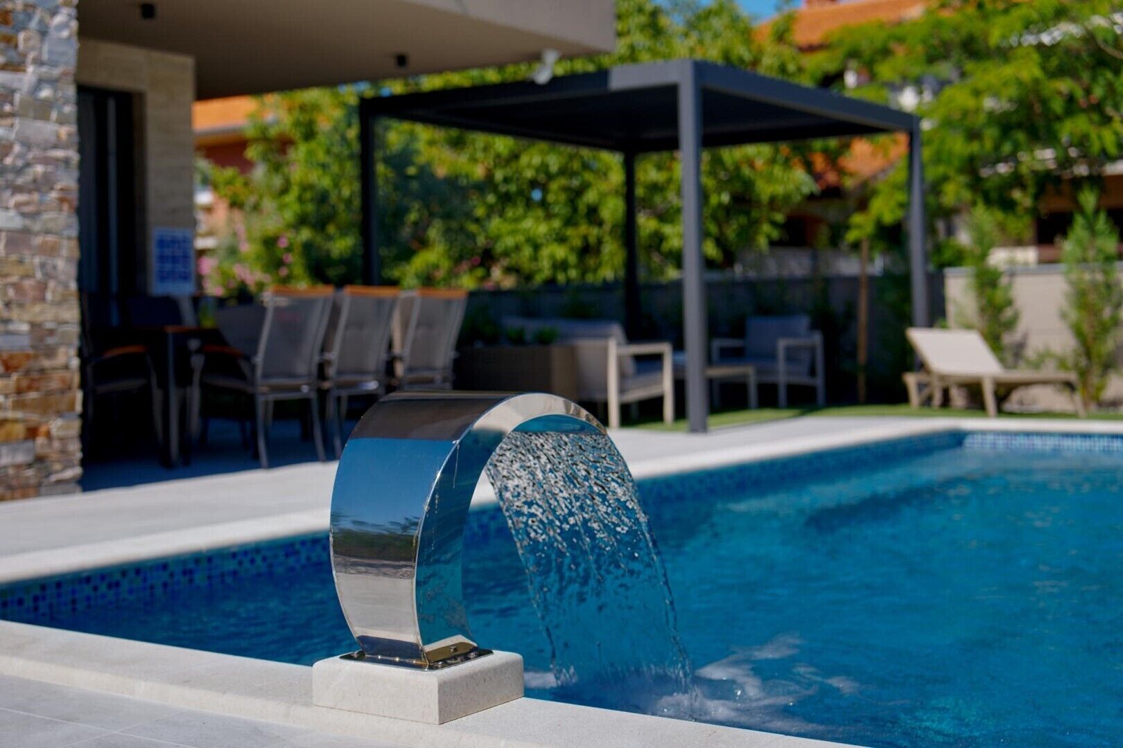 Une piscine extérieure moderne agrémentée d’un élément décoratif aquatique, entourée de chaises longues et d’un espace salon abrité.